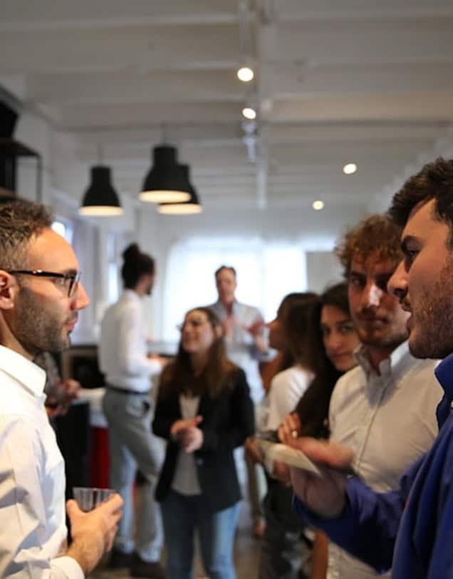 Group of men in the foreground at a networking event, with men and women in the background, engaging in professional connections and business discussions